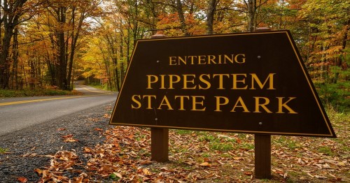 Sign for Pipestem State Park with a scenic road and autumn foliage in the background.