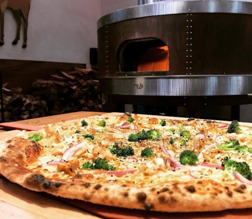 A close-up of a freshly baked pizza topped with broccoli and red onions, with a wood-fired oven in the background.