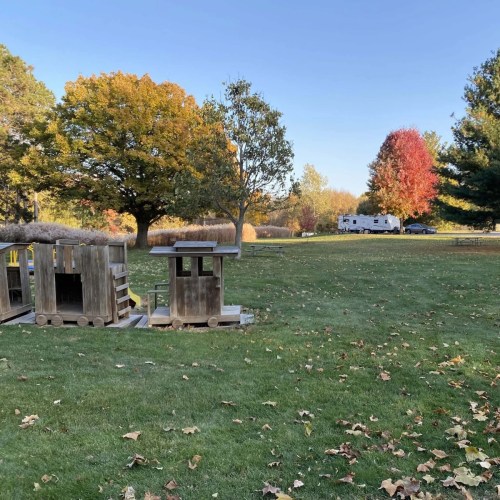 A grassy area with wooden play structures, colorful autumn trees, and an RV in the background under a clear blue sky.
