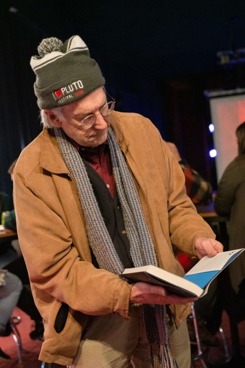 An older man in a beanie and scarf reads a book while standing in a casual indoor setting.