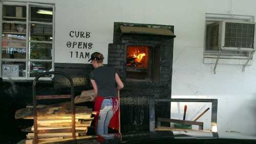 A person in an apron prepares wood near a brick oven with flames, next to a sign that says "CURB OPENS 11AM."