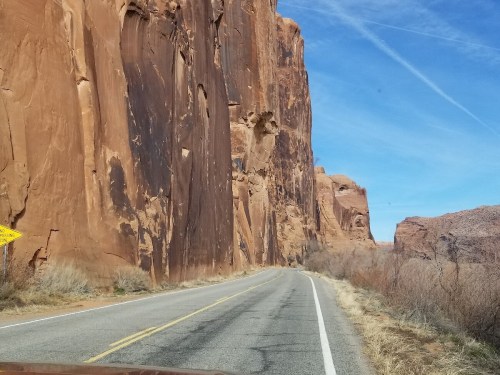 A winding road alongside towering red rock cliffs under a clear blue sky.