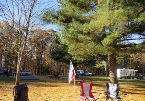 A campsite with two chairs, an American flag, and trees, surrounded by autumn foliage and parked RVs in the background.
