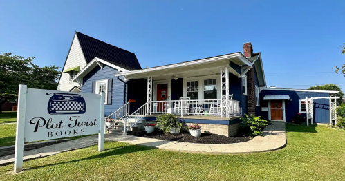 A charming blue house with a sign reading "Plot Twist Books" in front, featuring a porch and well-kept landscaping.