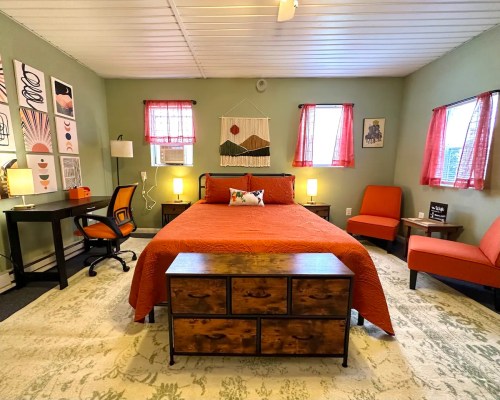 Cozy bedroom with a large bed, two chairs, a desk, and warm lighting, featuring colorful decor and a patterned rug.