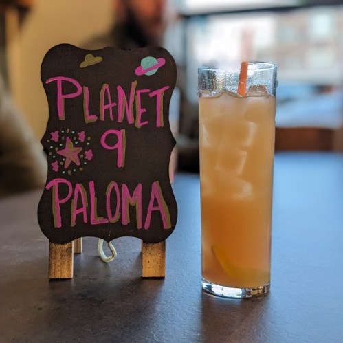 A chalkboard sign reads "Planet 9 Paloma" next to a tall glass of a light-colored cocktail with ice and a straw.