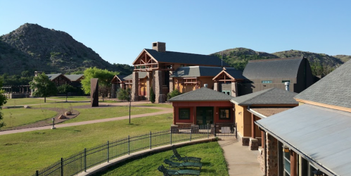 A scenic view of a modern building complex surrounded by green grass and mountains under a clear blue sky.