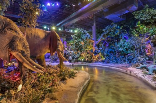 A lifelike elephant statue near a winding stream, surrounded by vibrant, colorful foliage in a themed indoor setting.