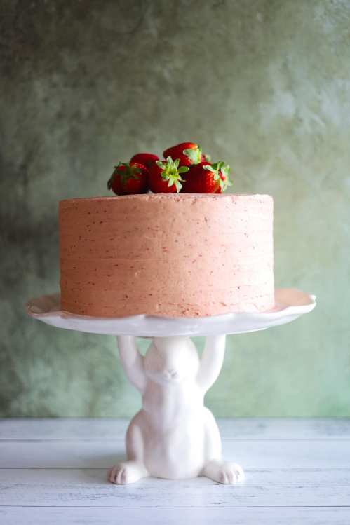 A pink frosted cake topped with fresh strawberries, displayed on a white rabbit-shaped cake stand.