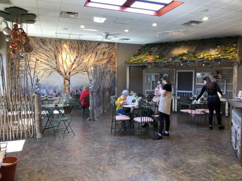 A cozy café interior with rustic decor, featuring a large tree mural, green chairs, and patrons enjoying their meals.