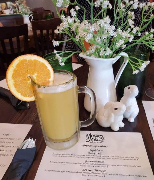 A glass of orange drink with a slice of orange, alongside a white pitcher and decorative bunnies on a table.