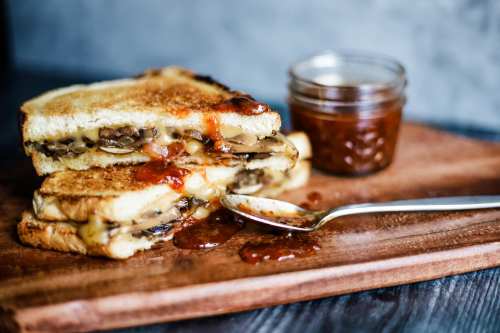 A stacked grilled cheese sandwich with mushrooms, oozing cheese, served with a jar of sauce on a wooden board.