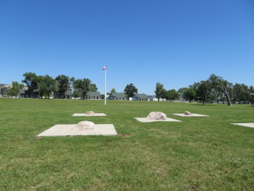 A grassy field with several concrete slabs and rocks, featuring a flagpole and buildings in the background under a clear blue sky.