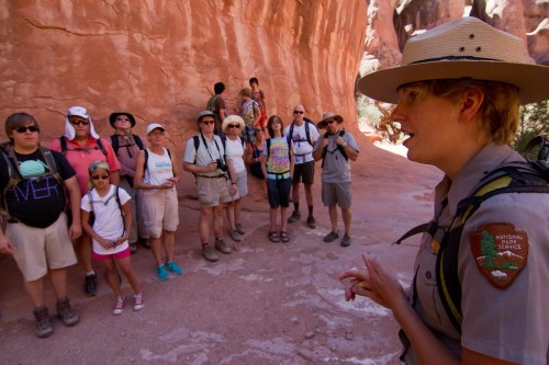 A park ranger speaks to a diverse group of hikers in a red rock canyon. Sunlight illuminates the scene.