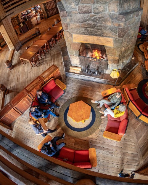 A cozy lodge interior with a stone fireplace, surrounded by red seating and wooden decor, where people are gathered.