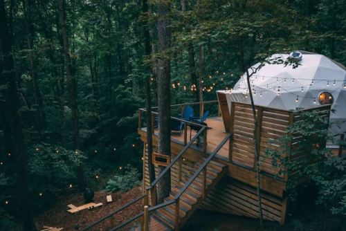 A geodesic dome cabin on a wooden deck surrounded by trees, with string lights and outdoor seating.