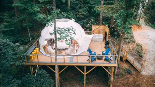 A geodesic dome cabin on a wooden deck in a forest, surrounded by trees and outdoor seating.