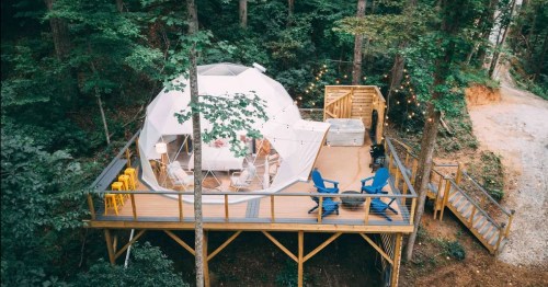 A cozy geodesic dome cabin on a wooden deck surrounded by trees, with outdoor seating and string lights.