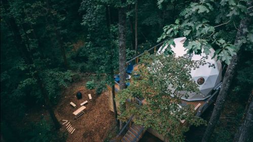 Aerial view of a geodesic dome nestled in a forest, surrounded by trees and a cozy outdoor seating area.