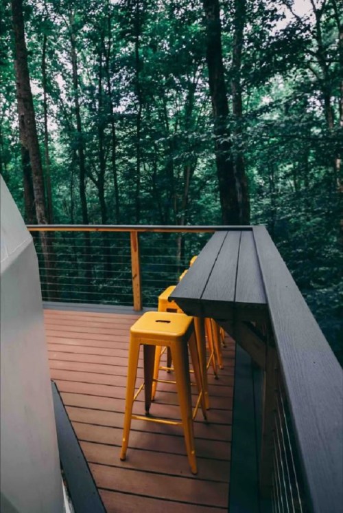 A wooden deck in a forest with three yellow stools overlooking lush greenery.