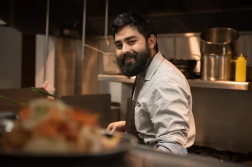 A smiling chef with a beard stands in a kitchen, preparing food with ingredients in the foreground.
