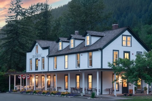A charming white hotel with multiple windows, surrounded by trees and a scenic mountain backdrop at sunset.