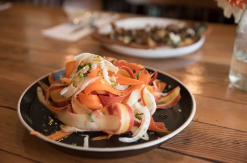 A colorful salad of carrot and beet ribbons topped with herbs, served on a black plate on a wooden table.