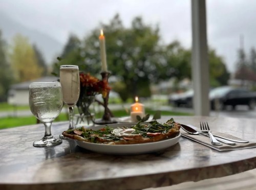 A cozy table setting with a plate of pizza, a glass of water, and a candle, set against a rainy outdoor backdrop.