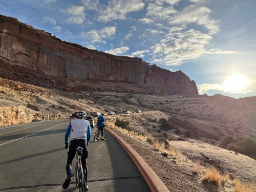Two cyclists ride along a winding road with rocky cliffs and a sunset in the background.