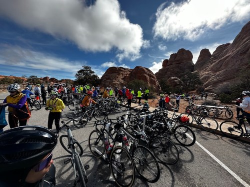 A large group of cyclists in bright gear gathers near their bikes, with rocky formations and a cloudy sky in the background.