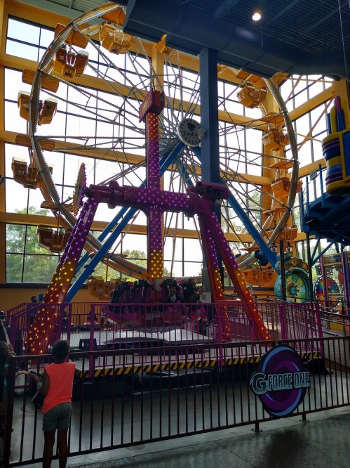 A colorful indoor amusement ride featuring a large Ferris wheel and a spinning attraction, with a child watching nearby.
