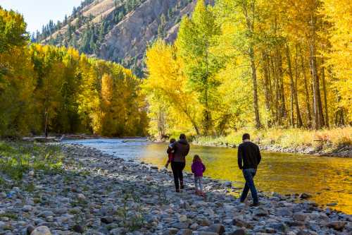 A family walks along a rocky riverbank surrounded by vibrant autumn trees and mountains in the background.