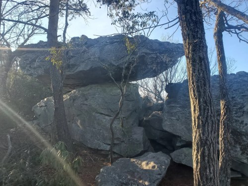 A large boulder balanced on top of a rock formation, surrounded by trees and sunlight filtering through.