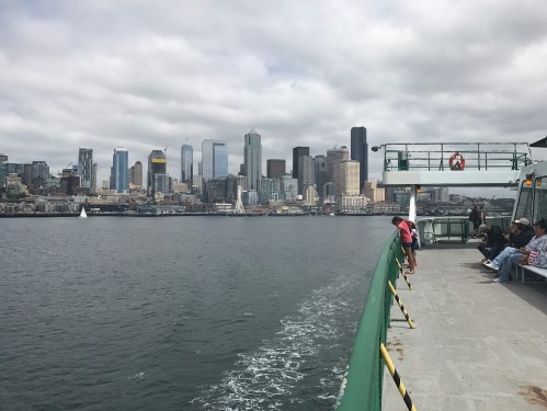 View from a ferry with passengers on board, overlooking a cloudy Seattle skyline across the water.