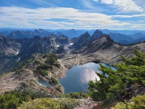 A scenic mountain landscape featuring rugged peaks, a shimmering lake, and a clear blue sky with scattered clouds.