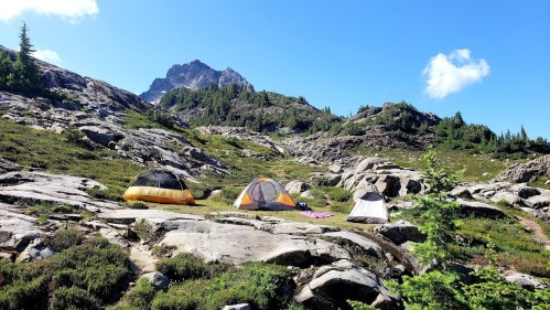 A scenic campsite with several tents set among rocky terrain and lush greenery, under a clear blue sky.