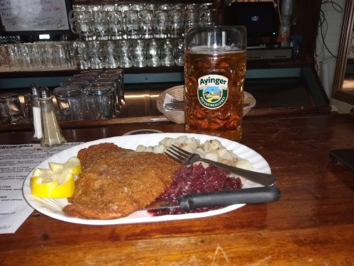 A plate with breaded fish, lemon, red cabbage, and potatoes, alongside a large beer on a bar counter.