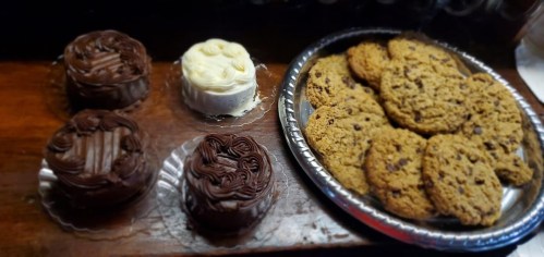 A wooden surface with two chocolate cakes, a white frosted cake, and a plate of chocolate chip cookies.