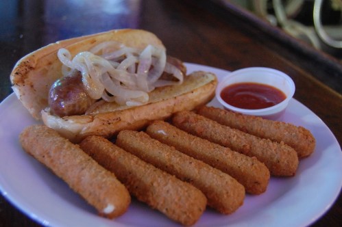 A plate featuring a sausage sandwich with onions and a side of crispy mozzarella sticks and marinara sauce.