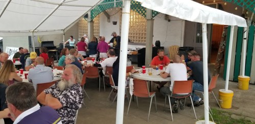 A group of people dining under a tent, with tables set up and food being prepared in the background.