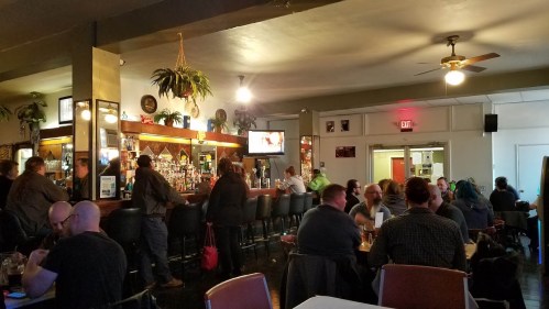 A lively bar scene with patrons at the counter and tables, plants hanging, and a TV in the background.