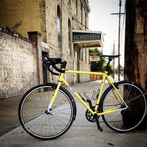 A yellow bicycle parked on a sidewalk in front of a brick building with a sign that reads "Sachsenheim."