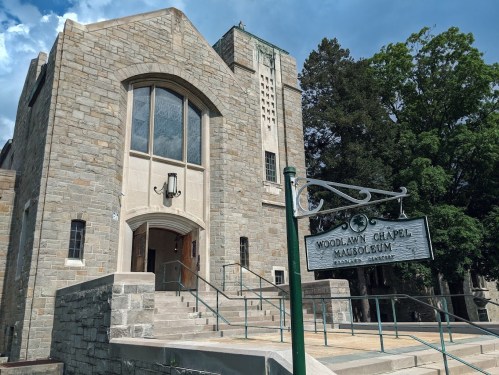 Stone building with large windows and a sign reading "Woodlawn Chapel Mausoleum" under a blue sky with clouds.