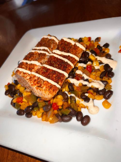 A plate of seasoned fish fillets on a bed of black beans and corn, drizzled with a creamy sauce.