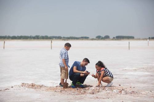 Two children and an adult explore a dry, salt flat, examining the ground under a clear sky.