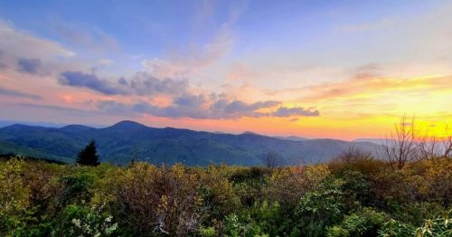 A scenic view of rolling mountains under a colorful sunset, with lush greenery in the foreground.