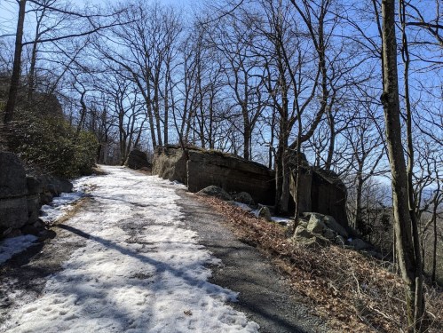 A snowy path winds through a wooded area with bare trees and rocky outcrops under a clear blue sky.
