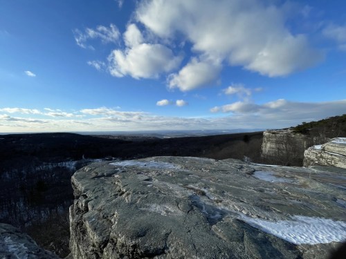 A rocky cliff edge overlooks a vast landscape under a blue sky with fluffy clouds. Snow is visible on the rocks.