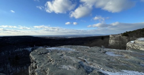 A panoramic view from a rocky cliff, showcasing a vast landscape of trees and distant hills under a blue sky with clouds.