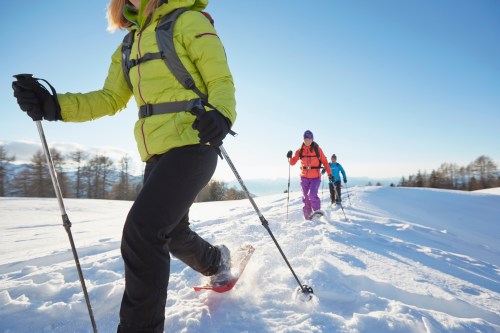 Three people snowshoeing on a snowy landscape under a clear blue sky, wearing colorful winter gear.